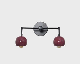 May include: A wall-mounted light fixture featuring a matte black circular base and a horizontal metal arm. Two adjustable stems extend from the center, each holding a glossy, deep burgundy dome-shaped shade with a small, warm-toned light bulb visible.