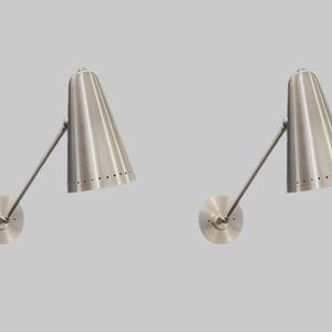 Op de afbeelding: Twee wandkandelaars van geborsteld nikkel. Elke wandkandelaar heeft een conische kap met een rij kleine gaatjes aan de onderkant, een gebogen arm en een ronde wandbevestiging. De lampen staan tegen een lichtgrijze achtergrond.