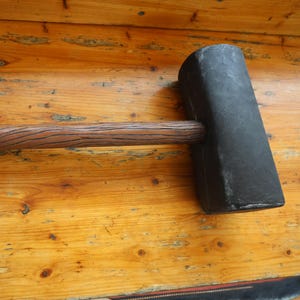May include: A large, black sledgehammer with a textured wooden handle. The hammerhead is rectangular and dark, contrasting with the brown handle. The handle has a wood grain pattern. The tool rests on a wooden surface.
