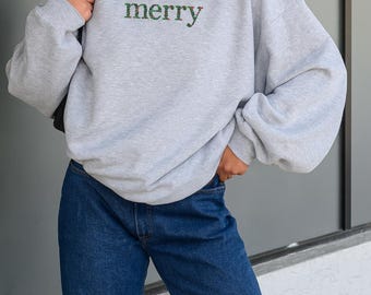 May include: A light gray sweatshirt with the word "merry" embroidered in green and red thread across the chest. The sweatshirt has a crew neck and long sleeves. The model is wearing blue jeans.