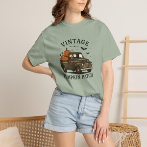 Puede incluir: Una camiseta de manga corta color verde salvia con un estampado gráfico de un camión antiguo verde oscuro que transporta calabazas naranjas. El texto VINTAGE PUMPKIN PATCH está impreso en negro sobre y bajo el camión, junto a pequeñas siluetas de murciélagos.