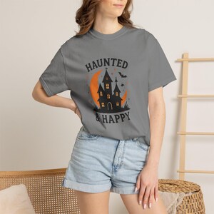 Puede incluir: Una camiseta gris carbón de manga corta con un estampado gráfico de la silueta de una casa encantada negra sobre una luna creciente naranja. El texto "HAUNTED & HAPPY" está impreso en negro sobre y bajo la casa, junto a estrellas y un murciélago.