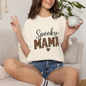 Puede incluir: Una camiseta de manga corta color crema con el texto cursivo negro Spooky sobre la palabra MAMA en un patrón de leopardo marrón y negro, con un pequeño icono de corazón con estampado de leopardo centrado bajo el texto en la parte frontal.