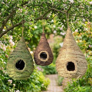 May include: Three hanging birdhouses in a garden setting. The birdhouses are teardrop-shaped and made of woven natural materials in shades of green, brown, and tan. Each birdhouse has a circular opening and is suspended from a tree branch.