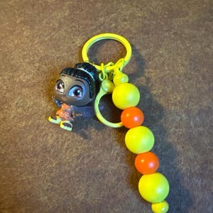 May include: A yellow keychain with a small figurine of a person with dark hair and brown eyes. The keychain has a series of yellow and orange beads. The figurine is wearing an orange and brown outfit.