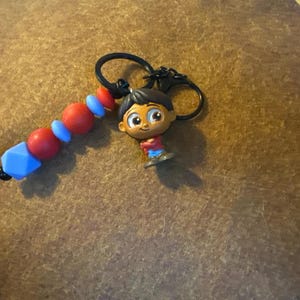 Puede incluir: Un llavero con una pequeña figura de un personaje de dibujos animados con cabello oscuro, que lleva una camisa roja y pantalones azules. El llavero también incluye una cadena de cuentas rojas, azules y una cuenta geométrica azul.