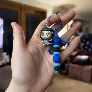 May include: A keychain featuring a small figurine of a character with black hair and a blue dress. The keychain has a blue ring and a beaded design in shades of blue and black. The figurine has large eyes and is attached to the keychain.