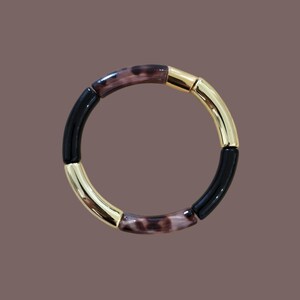 Peut inclure: Un bracelet circulaire avec des segments alternés noirs, dorés et marron à motifs. Les segments dorés ont une finition brillante, tandis que les sections noires et marron ont un aspect lisse. Le bracelet est posé sur un fond marron uni.