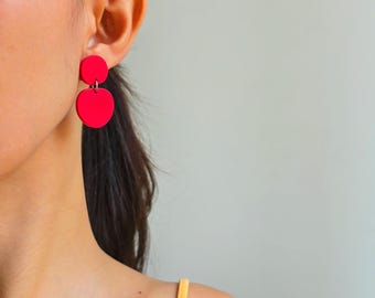 May include: Bright red, two-piece earrings. The top piece is a small circle, and the bottom piece is a heart shape. The earrings are worn on a person's ear, with the background a neutral gray.