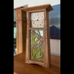 May include: A wooden mantel clock with a square face and a decorative dragonfly panel. The clock face has black numerals and hands. The dragonfly panel features colorful dragonflies on a green background. The clock is made of light brown wood.