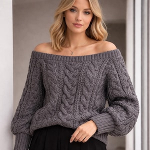 Op de afbeelding: Een grijze off-shoulder trui met een kabelgebreid patroon. De trui heeft lange mouwen met geribbelde manchetten en een brede halslijn. Het materiaal lijkt een zachte, warme brei te zijn, perfect voor koeler weer.