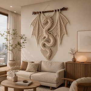 May include: A large, intricate macrame wall hanging featuring a central dragon figure with expansive, detailed wings. The piece is crafted from cream-colored cord and suspended from a natural wooden branch, creating a textured, sculptural focal point.