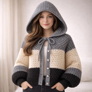 May include: A hooded crochet jacket in gray, cream, and black. The jacket features a drawstring closure and silver buttons. The sleeves are a darker gray and black, with a matching hood. The jacket is a cozy, handmade garment.