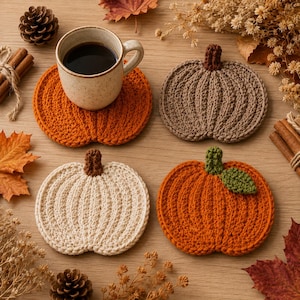 May include: A set of four crocheted pumpkin-shaped coasters in orange, beige, and brown. One orange coaster supports a speckled ceramic mug filled with dark coffee. Each coaster features a ribbed texture and a small stem, with one featuring a green leaf.
