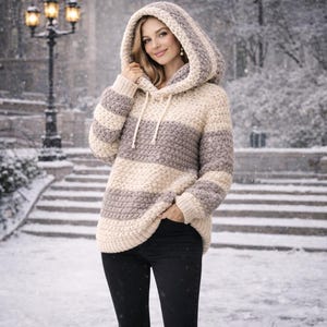 May include: A cream, grey, and white striped hooded sweater. The sweater features a drawstring hood and ribbed cuffs. The person in the image is wearing black trousers and is outside in a snowy scene.