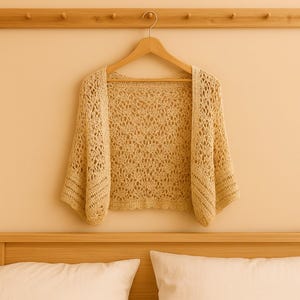 Könnte beinhalten: Ein beigefarbener, offener Häkel-Bolero mit kurzen Ärmeln hängt an einem Holzbügel. Das zarte, offene Gewebe des Kleidungsstücks ist sichtbar und hängt an einer hellfarbigen Wand. Der Bolero hat eine helle, neutrale Farbe.