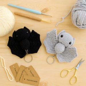 May include: Two crocheted bat keychains, one black and one gray, with key rings. The bats have detailed wings and small eyes. Crafting tools, including crochet hooks, yarn, and scissors, are arranged around the bats. Small tags read "handmade".