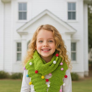 May include: A young girl with blonde curly hair smiles, wearing a green crocheted scarf adorned with colorful pom-poms. The scarf is wrapped around her neck, and she is wearing a white long-sleeved shirt. A white house is in the background.
