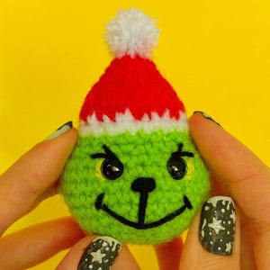 May include: A crocheted green character with black eyes and a smiling mouth, wearing a red and white Santa hat. The character is round and has a textured appearance, perfect for holiday decor.