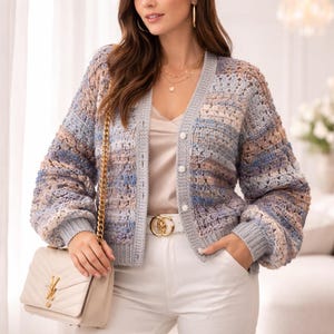 May include: A multi-colored, open-weave cardigan in shades of blue, gray, and tan. The sweater has a V-neck, button closure, and long sleeves. A cream-colored handbag with a gold chain strap is visible. The cardigan is part of a clothing collection.