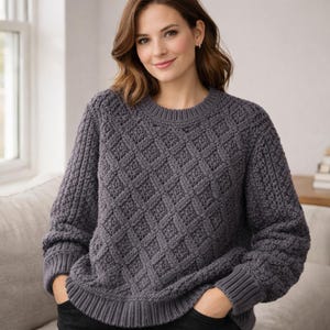 Könnte beinhalten: Ein grauer Pullover mit Rautenmuster, Rundhalsausschnitt und langen Ärmeln. Die Strickstruktur ist sichtbar, und der Pullover scheint aus einem weichen, warmen Material zu sein. Die Person trägt eine schwarze Hose.