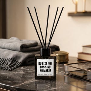 Op de afbeelding: Een vierkante, donkere glazen diffuser met zwarte stokjes. De fles heeft een zwarte dop en een wit etiket met zwarte tekst: "DU BIST 40? DAS SIND 80 MARK!". De diffuser staat op een marmeren oppervlak.
