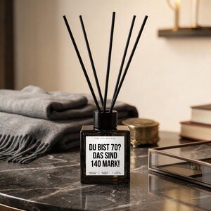 Op de afbeelding: Een zwarte vierkante glazen diffuserfles met zwarte stokjes. Het etiket luidt "DU BIST 70? DAS SIND 140 MARK!". De diffuser staat op een marmeren oppervlak, met een stapel grijze dekens op de achtergrond.