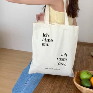 Könnte beinhalten: Cremefarbene Tragetasche mit schwarzem Text: "ich atme ein. ich raste aus." und der Website-Adresse "www.attitudecandles.de". Die Tasche hat ein schlichtes Design mit zwei Schulterriemen.