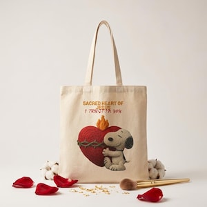 May include: Beige tote bag featuring a graphic of Snoopy hugging a red heart with a crown of thorns. The heart has a flame on top. Text on the bag reads "Sacred Heart of Jesus, I Trust in You."