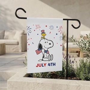Może przedstawiać: Biała flaga ogrodowa z Snoopy'm i Woodstockiem świętującymi 4 lipca. Snoopy trzyma amerykańską flagę i nosi patriotyczną chustę. Flaga ma czerwone, białe i niebieskie fajerwerki oraz napis "JULY 4TH".