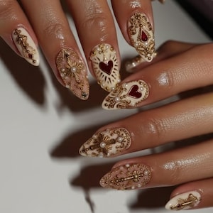 May include: Close-up of elaborately decorated nail art. The nails have a cream base with gold embellishments, including crosses, hearts, and floral patterns. Small pearls and red accents enhance the design. The nails are long and pointed.