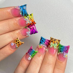 May include: A close-up of a hand with long, colorful, and decorated acrylic nails. The nails feature a variety of designs, including animal prints, floral accents, and rhinestones. The nail beds are painted a light pink color.