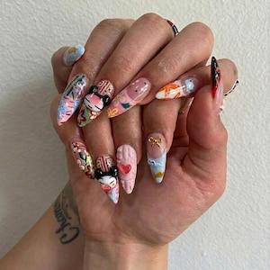 May include: A hand with long, almond-shaped nails, each adorned with unique designs. The nails feature a variety of colors, including pink, blue, and orange, with intricate patterns, cartoon characters, and 3D embellishments. Some nails have a clear base with painted designs.