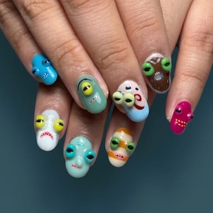 May include: A hand with a manicure featuring whimsical, 3D monster faces on each nail. The nails are painted in various colors, including blue, green, pink, and brown, with detailed facial features like eyes and mouths.