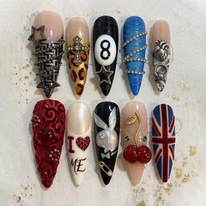 May include: A collection of ten long, almond-shaped artificial nails with various designs. The designs include a star and chain, a cross, a leopard print, a number 8 ball, blue rhinestones, a heart, a red velvet design, the words "I ❤️ ME", a Playboy bunny, and a cherry.
