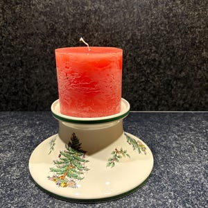 Op de afbeelding: Een rode pilaar kaars staat op een decoratieve keramische kandelaar. De kandelaar is wit met een groene rand en heeft een kerstboom- en cadeauontwerp. Een feestelijke vakantiedecoratie.