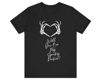 May include: Black t-shirt with a white skeleton hand heart design and the text "Will You Be My Spooky Pookie?" in a cursive font. The shirt is made of a soft material. Ideal for Halloween or spooky season.