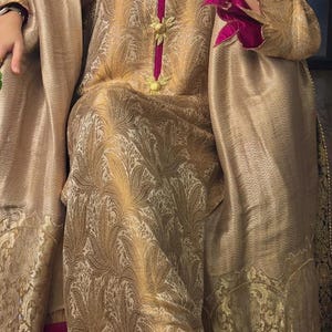 May include: A gold-colored, patterned ensemble featuring a long-sleeved top and matching pants. The outfit has a V-neckline with a magenta trim and decorative gold buttons. A matching shawl drapes over the shoulders, and the sleeves have magenta floral accents.