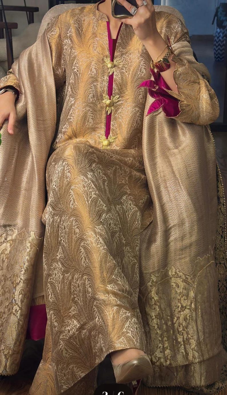 May include: A gold-colored, patterned ensemble featuring a long-sleeved top and matching pants. The outfit has a V-neckline with a magenta trim and decorative gold buttons. A matching shawl drapes over the shoulders, and the sleeves have magenta floral accents.