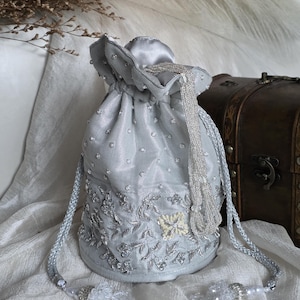 Puede incluir: Un bolso de satén azul claro con cordón, con intrincados bordados florales y pedrería dispersa. El bolso presenta una borla de cuentas y cordones trenzados. Un pequeño y elegante accesorio.