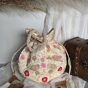 May include: Cream embroidered pouch bag with pink and red floral details. The bag has a pearl handle and tassel drawstrings. A small wooden chest is in the background.