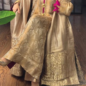 May include: A gold-colored ensemble featuring a long-sleeved top and wide-leg pants. The outfit has a floral pattern and is accented with pink trim and gold embellishments. The fabric has a subtle sheen, and the outfit is paired with gold-colored shoes.