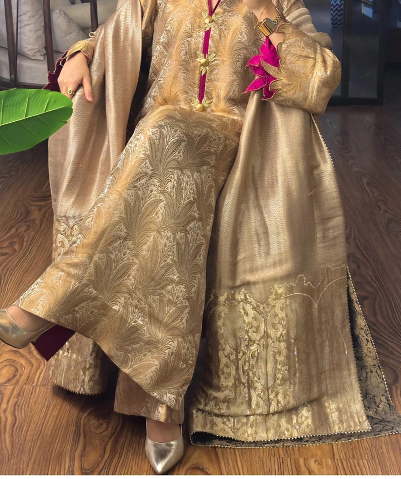 May include: A gold-colored ensemble featuring a long-sleeved top and wide-leg pants. The outfit has a floral pattern and is accented with pink trim and gold embellishments. The fabric has a subtle sheen, and the outfit is paired with gold-colored shoes.