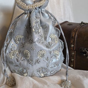 Puede incluir: Un bolso de satén azul claro con cordón, con intrincados bordados florales plateados y un ribete de perlas. El bolso presenta un borde festoneado y cordones con borlas plateadas. Un accesorio decorativo.