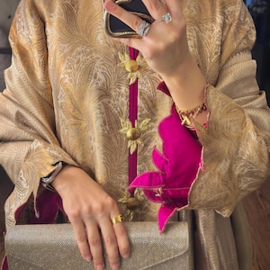 May include: A gold-colored, patterned jacket with a high collar and magenta trim. The jacket features gold floral embellishments and is paired with a silver clutch. The sleeves have a magenta floral design.