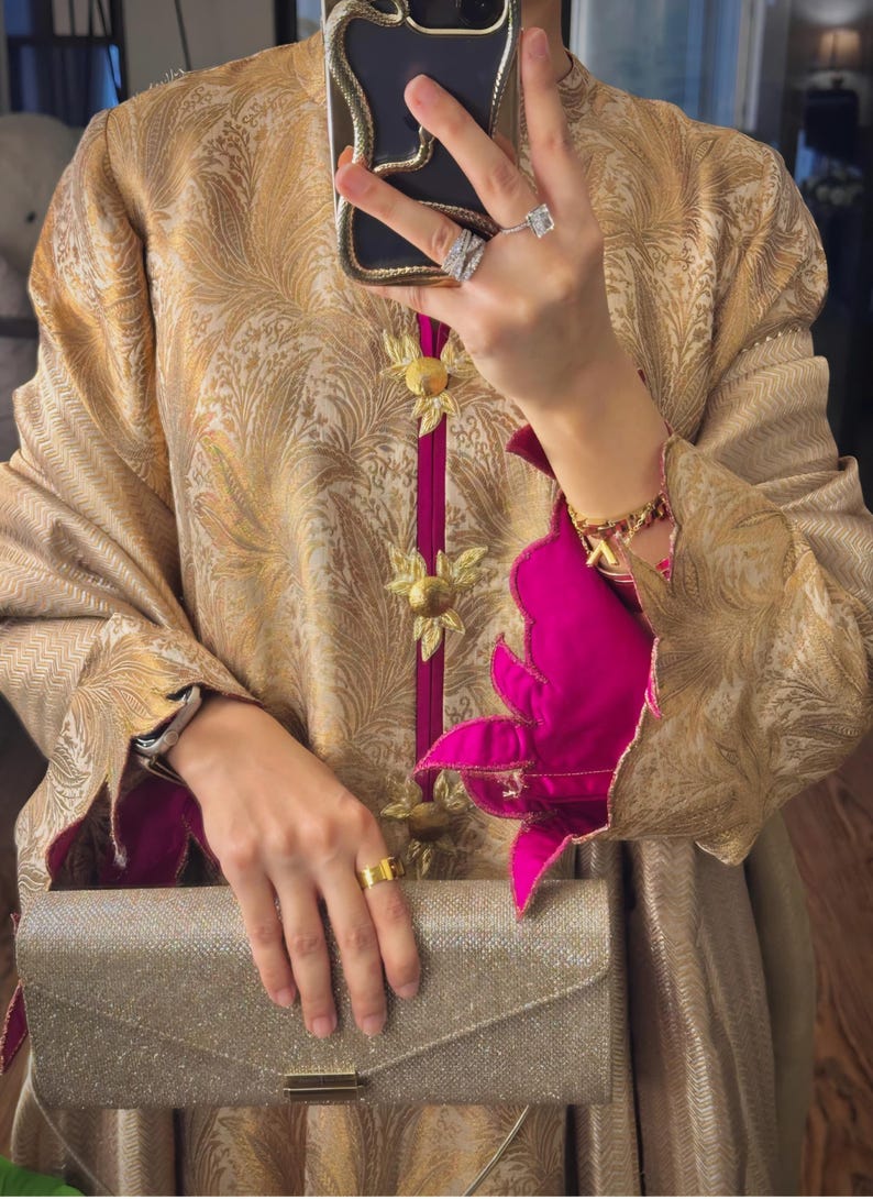 May include: A gold-colored, patterned jacket with a high collar and magenta trim. The jacket features gold floral embellishments and is paired with a silver clutch. The sleeves have a magenta floral design.