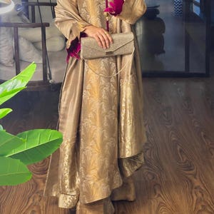 May include: A gold-colored formal outfit with a matching clutch. The outfit features a long, flowing coat with intricate patterns and a contrasting magenta lining. The clutch has a chain strap.