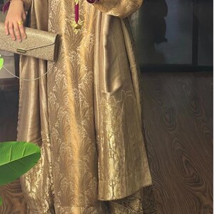 May include: A gold-colored, long-sleeved dress with an intricate floral pattern. The dress features a high neckline with a magenta trim and gold embellishments. A matching gold clutch is held in the hand. The dress is floor-length.