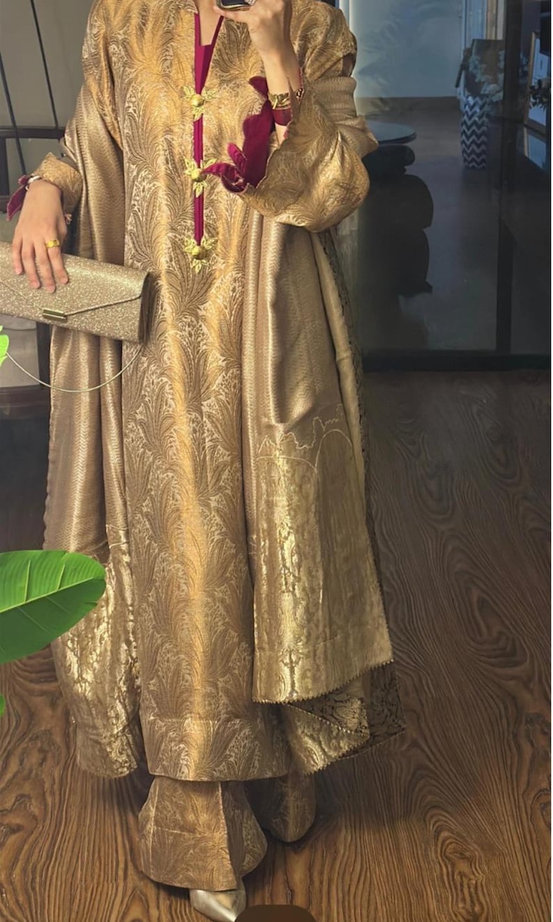 May include: A gold-colored, long-sleeved dress with an intricate floral pattern. The dress features a high neckline with a magenta trim and gold embellishments. A matching gold clutch is held in the hand. The dress is floor-length.