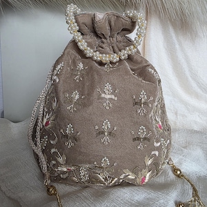 May include: A taupe velvet drawstring bag with detailed gold and white embroidery. The bag has a pearl-beaded handle and gold tassel detailing. The floral embroidery includes small pink accents, adding a touch of colour to the design.